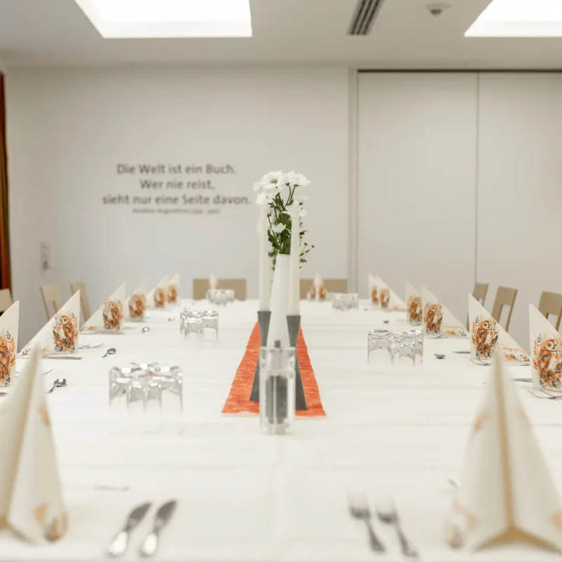 Heller Raum mit langer Festtafel und orangefarbenen Dekorationselementen.