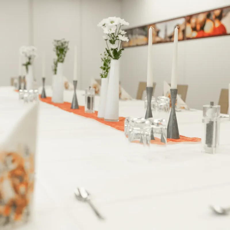 Lange Festtafel mit Blumenvasen, Gläsern und orangefarbenem Tischläufer.