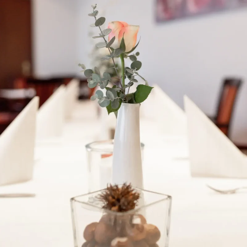 Rosafarbene Rose in weißer Vase zwischen gefalteten Servietten.