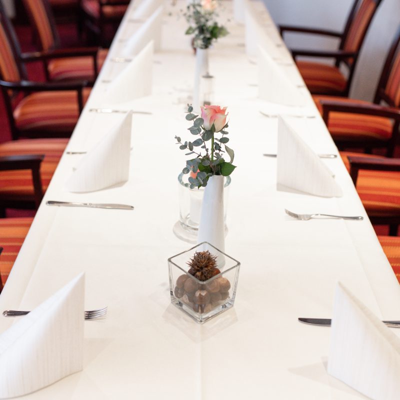 Lange, weiß gedeckte Festtafel mit Blumenvasen und gefalteten Servietten.