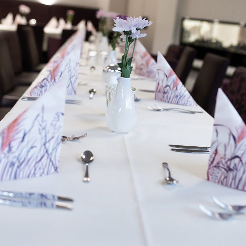 Lange gedeckte Tafel mit Blumen und lilafarben gemusterten Servietten.
