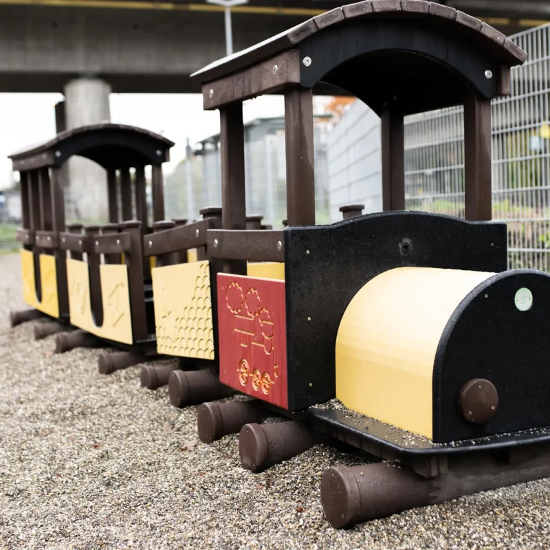 Spielzeuglokomotive aus Holz mit mehreren Waggons auf einem Kiesboden.