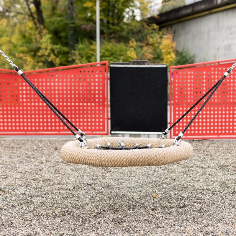 Runde Nestschaukel auf einem Spielplatz mit rotem Sicherheitszaun im Hintergrund.