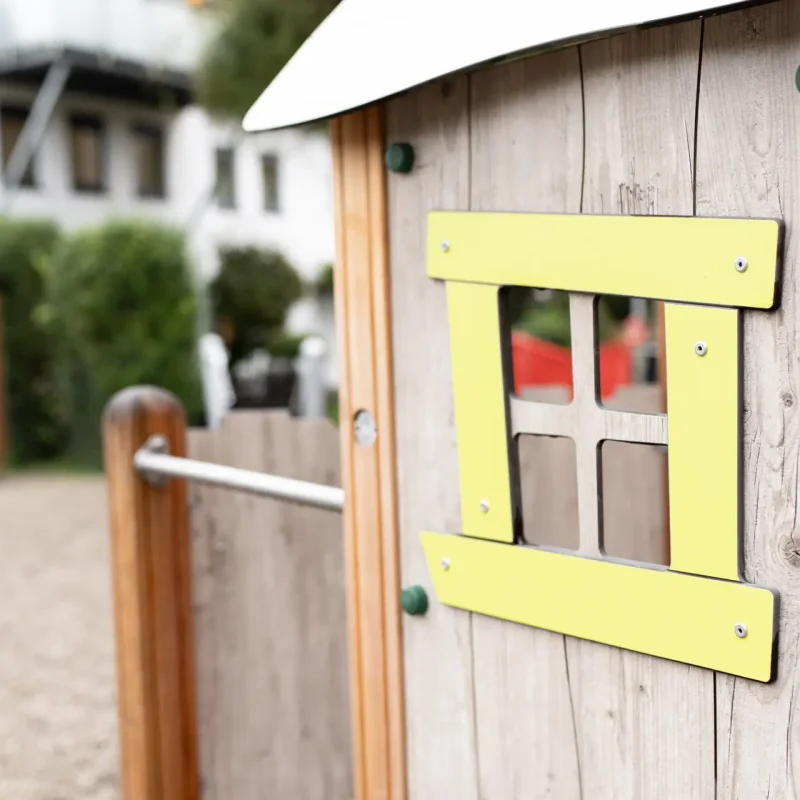 Detailansicht eines Spielhauses aus Holz mit gelbem Fensterrahmen.