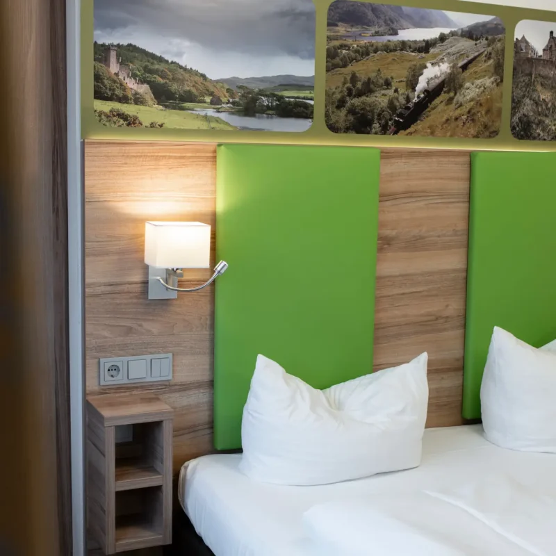 Hotelbett mit gr&uuml;nen Kopfteilpolstern, wei&szlig;en Kissen und Leselampe.