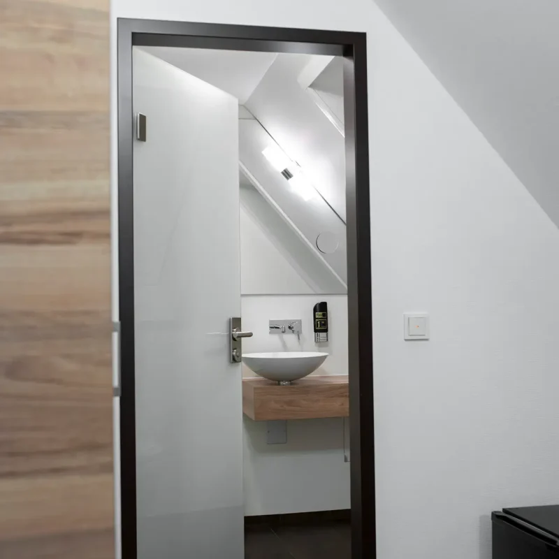 Ge&ouml;ffnete Hotelzimmert&uuml;r mit Blick in einen hellen Eingangsbereich und auf ein Waschbecken.