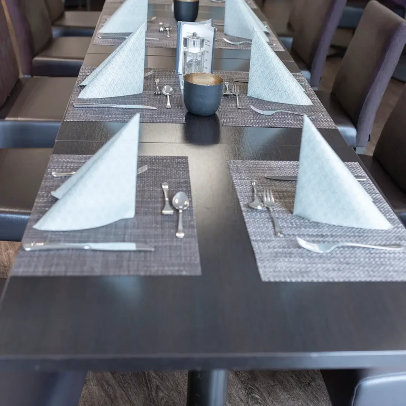 Lange Restauranttafel mit dunklen Tischsets, weißen Servietten und kleinen Tischdekorationen.