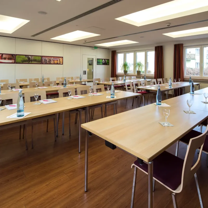 Tagungsraum mit U-förmig angeordneten Holztischen, Stühlen und großen Fenstern.