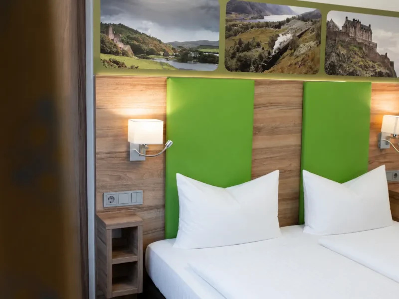 Hotelzimmer mit gr&uuml;nen Kopfteilpolstern, Holzverkleidung und Leselampe.