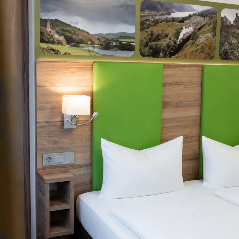 Hotelzimmer mit gr&uuml;nen Kopfteilpolstern, Holzverkleidung und Leselampe.