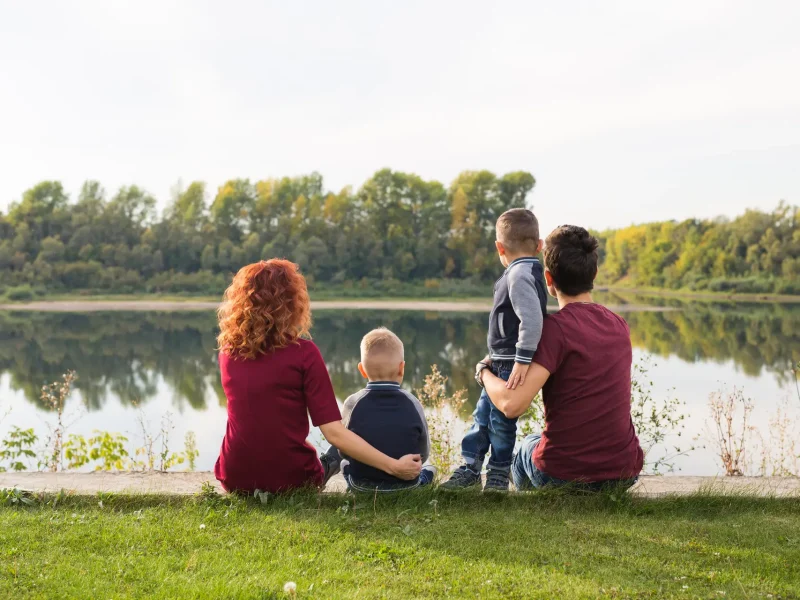 Familie mit zwei Kindern sitzt am Ufer eines Sees und blickt aufs Wasser.